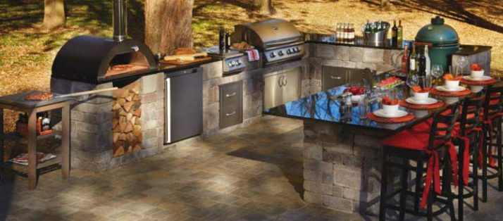 Outdoor kitchen appliances along with a glass dining table and red chairs under trees